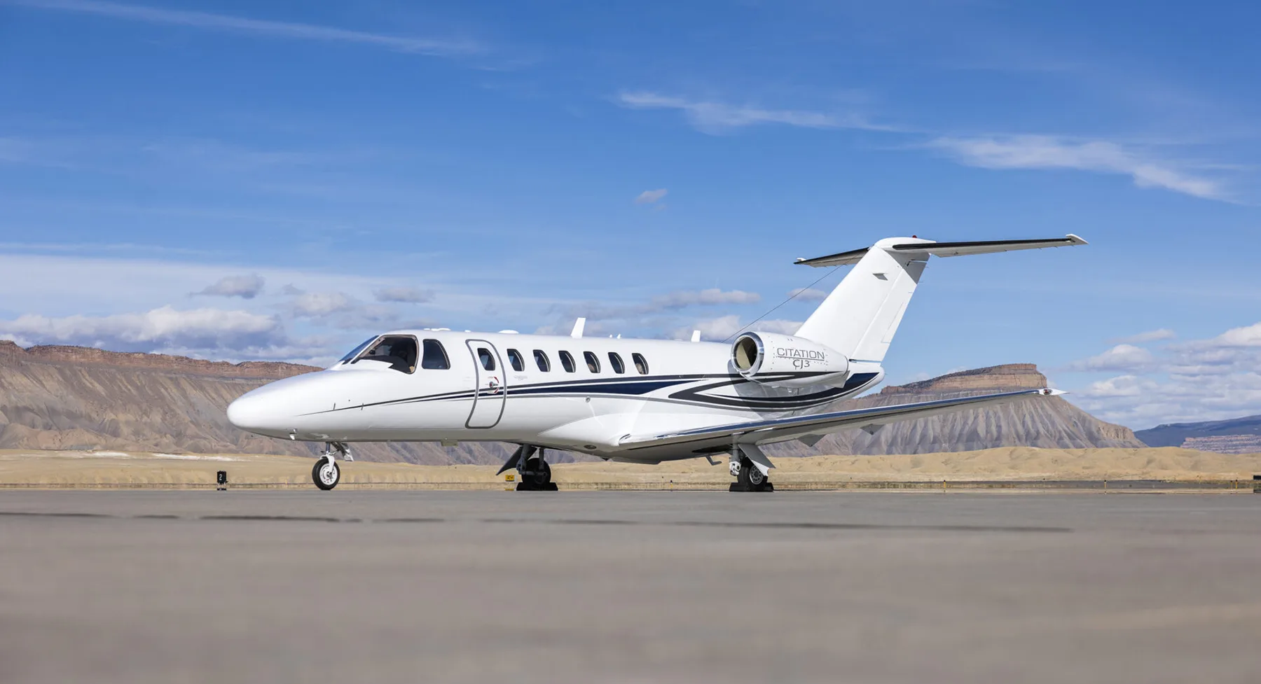 A white Cessna Citation CJ3 business jet with blue and black stripes is parked on an airfield with mountains in the background.