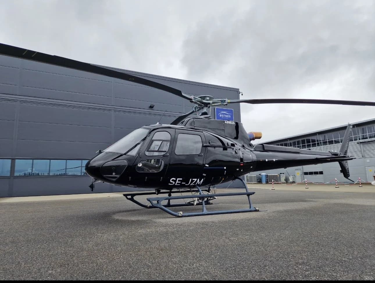 A black Airbus helicopter, registration SE-JZM, parked on an asphalt apron in front of a dark grey hangar under an overcast sky.
