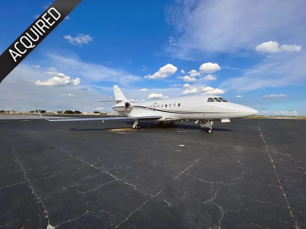 A white business jet with a dark stripe is parked on an asphalt tarmac under a blue sky with scattered clouds.