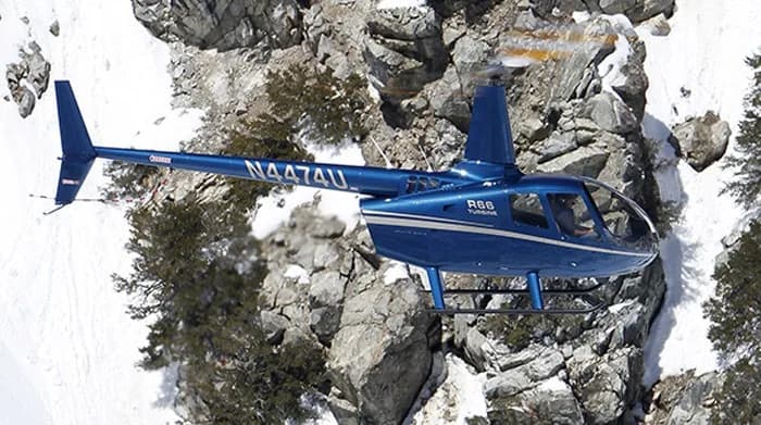 A blue Robinson R66 Turbine helicopter flies over a rocky, snowy landscape.