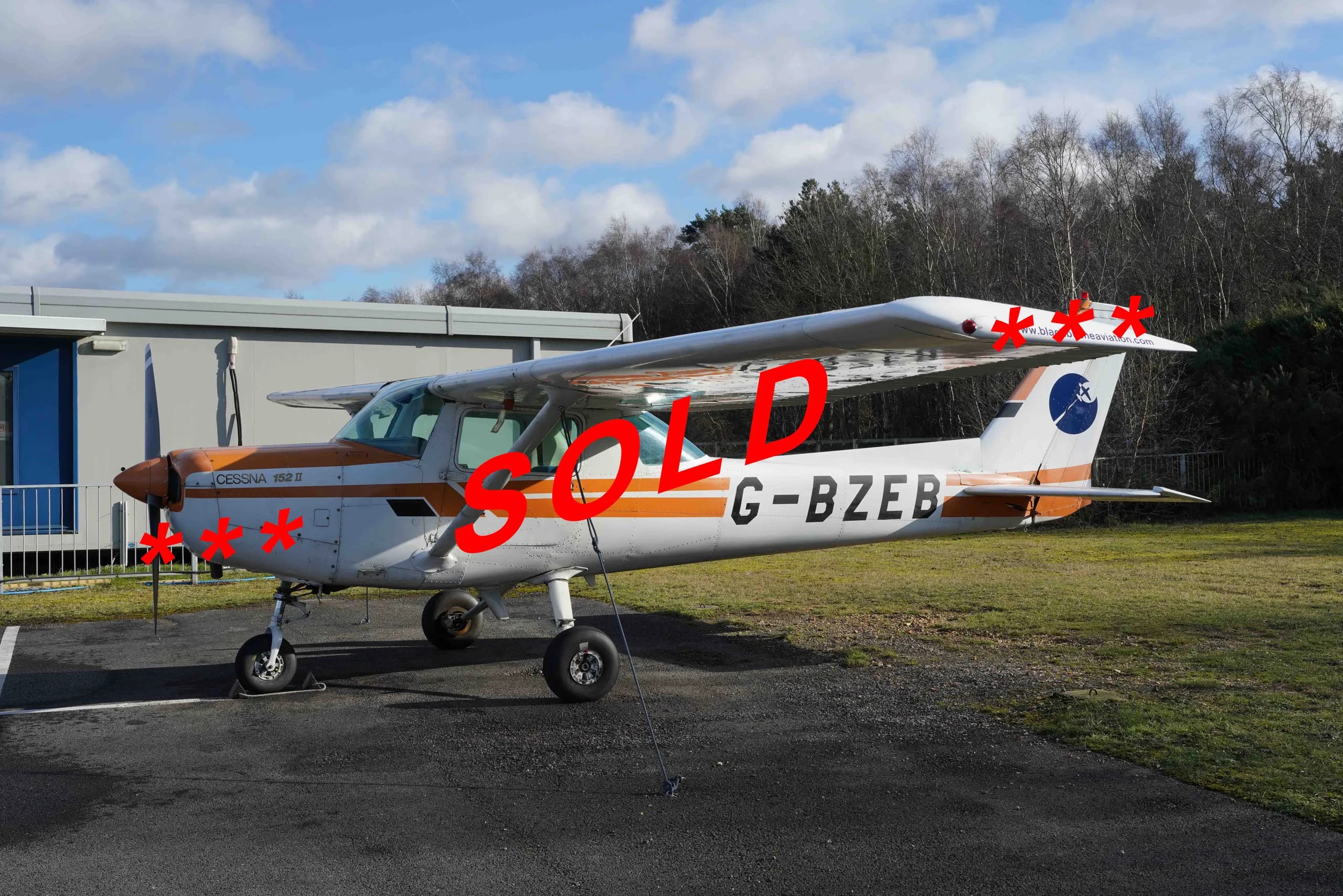 A white and orange Cessna 152 II aircraft, with registration G-BZEB, is parked on an asphalt ramp next to a grassy area, with a building and trees in the background.