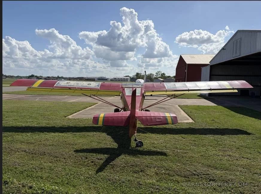 A rear view of a red and yellow ultralight aircraft parked on grass next to a hangar under a blue sky with clouds.