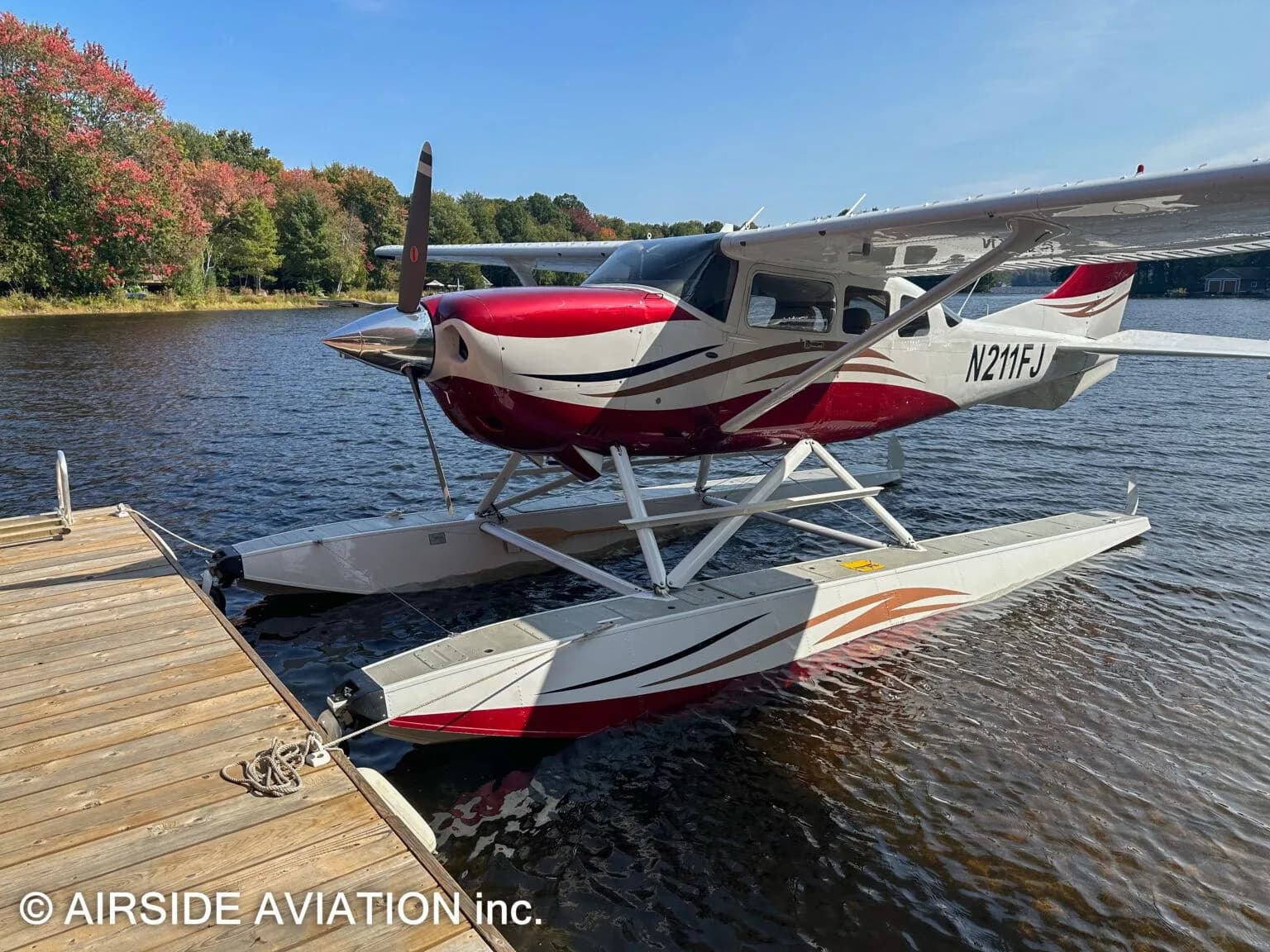 A red and white floatplane, N211FJ, is tied to a wooden dock on a lake surrounded by autumn foliage.