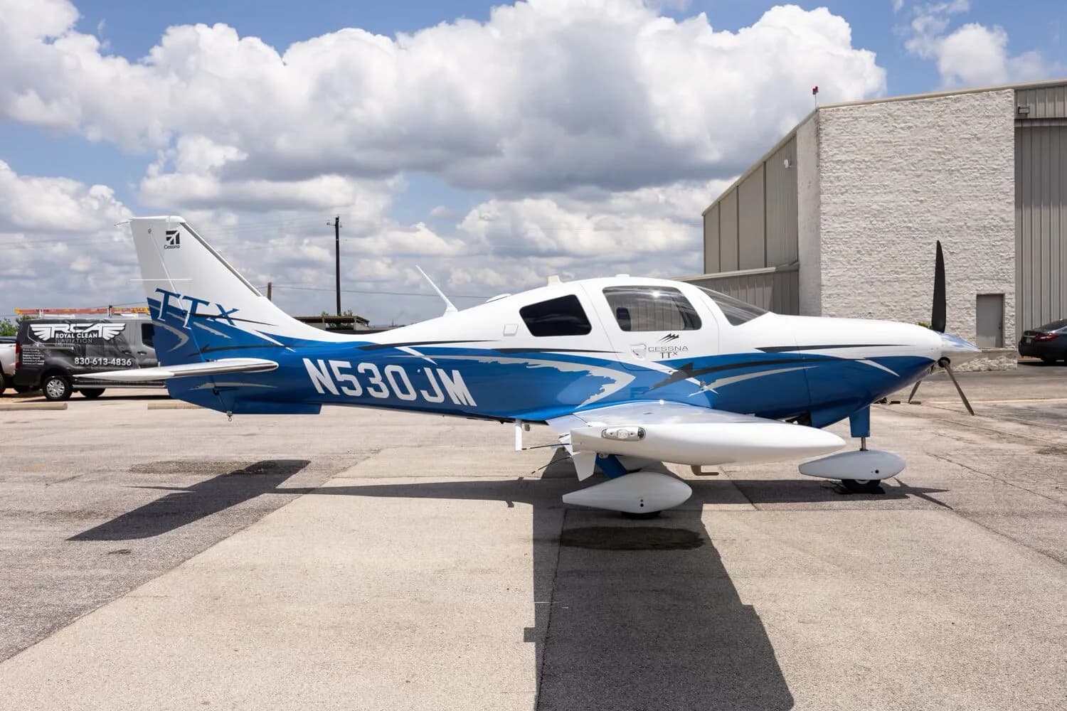A side view of the white and blue Cessna TTX N530JM parked on a tarmac next to a hangar.