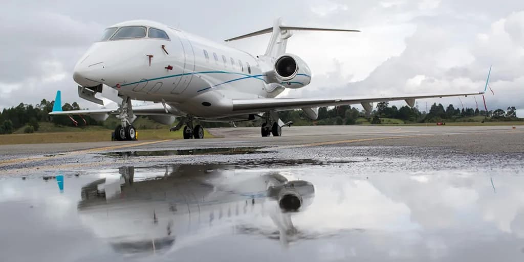A white business jet with blue and teal stripes is parked on a wet tarmac, reflecting in a puddle under a cloudy sky.