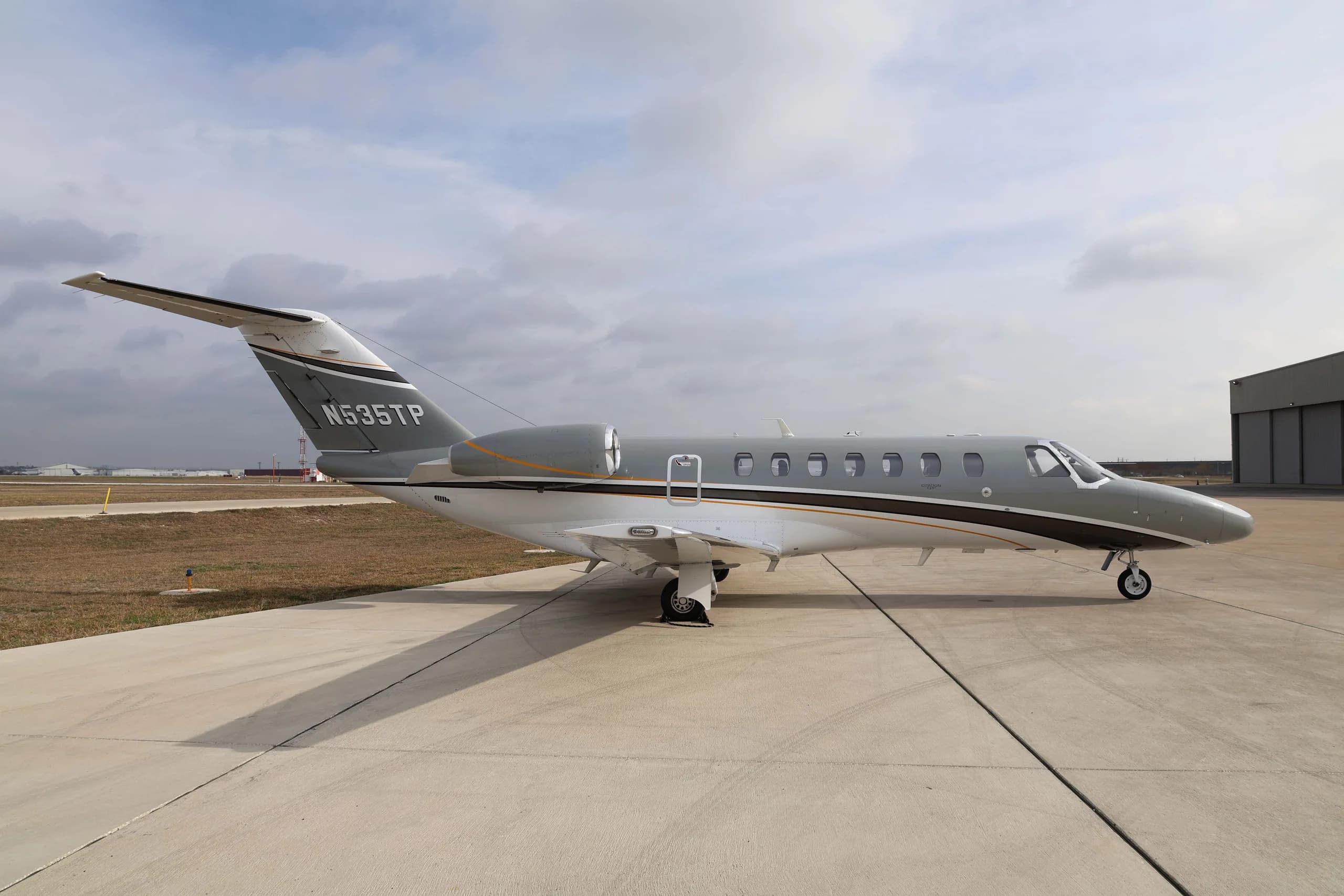 A side view of the Cessna Citation CJ3+ business jet, registration N535TP, with a grey and white paint scheme, parked on an airfield next to a hangar.