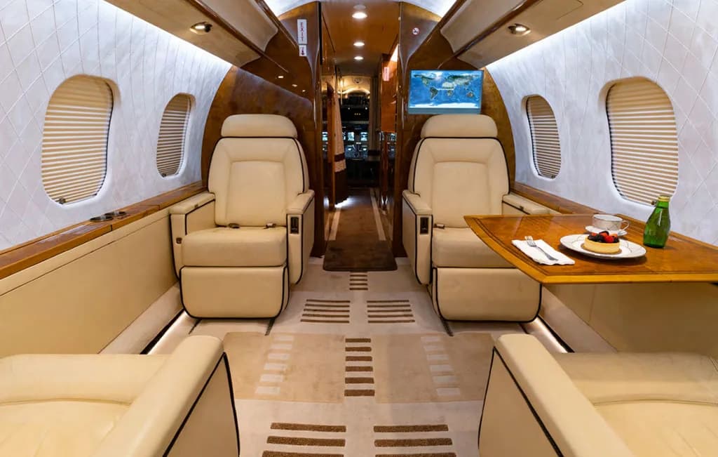 A view of the private jet cabin showing two beige seats facing a wooden table with a dessert and a map displayed on a screen.