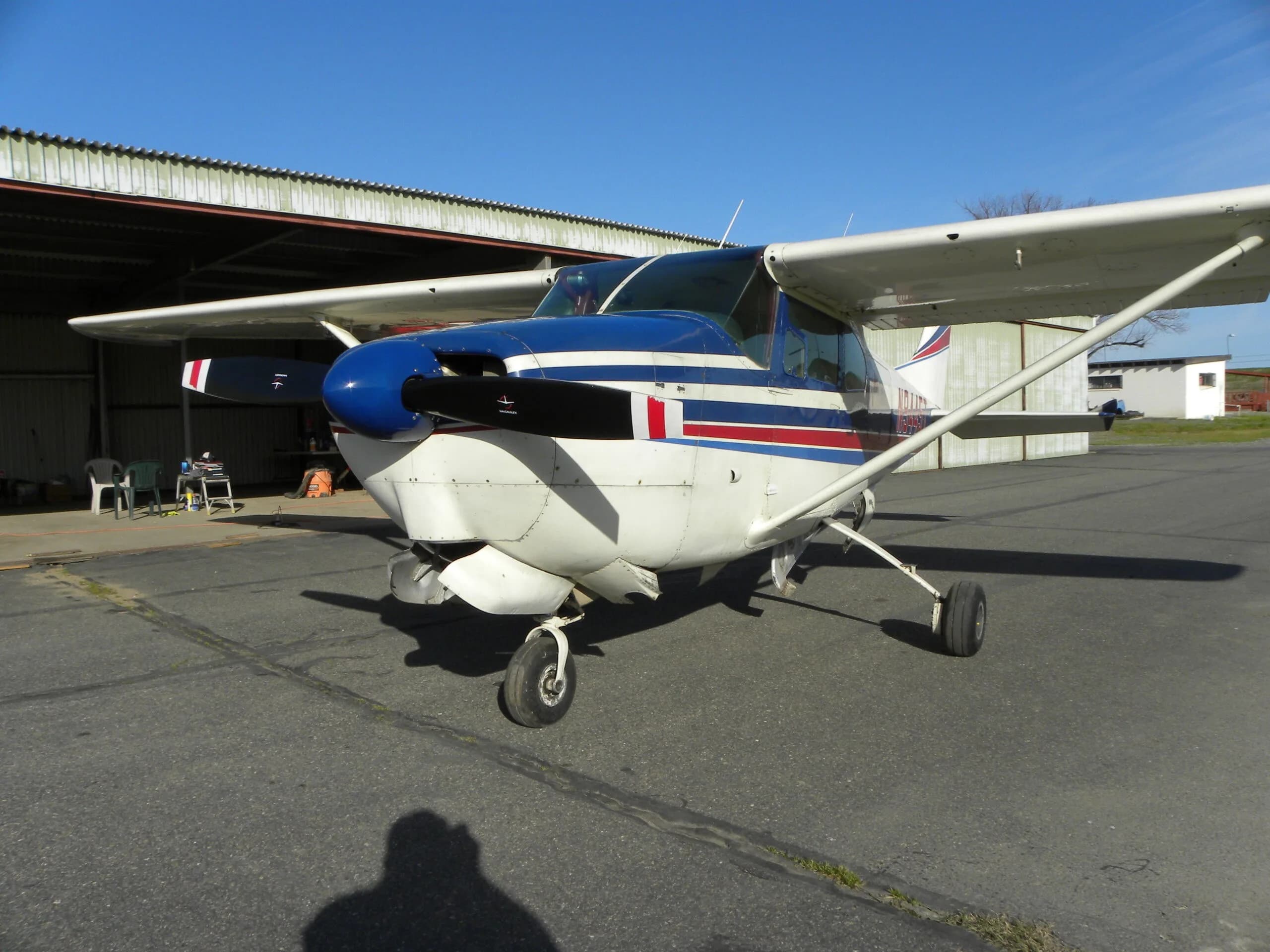 A white Cessna 182 with blue and red stripes parked on a tarmac in front of a hangar.