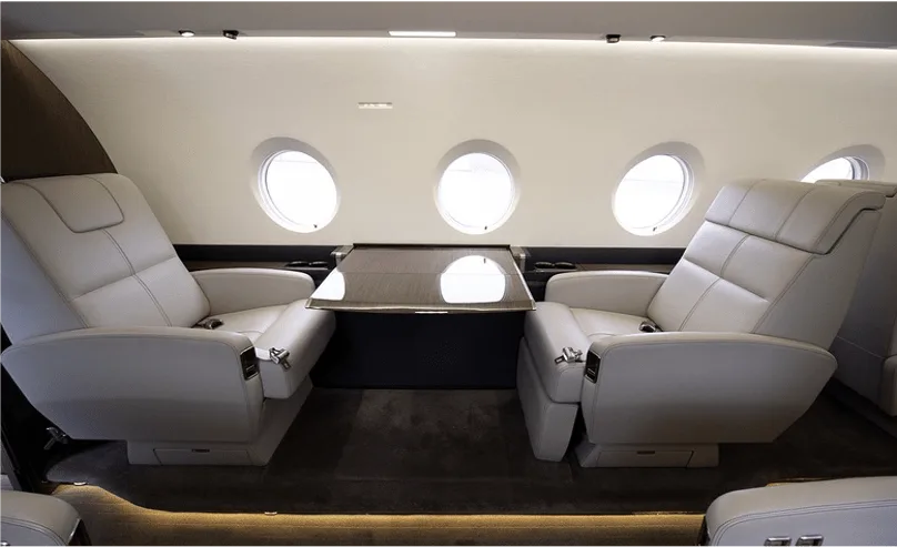 Two light leather seats facing each other with a small table in between, next to three oval windows in an aircraft cabin.