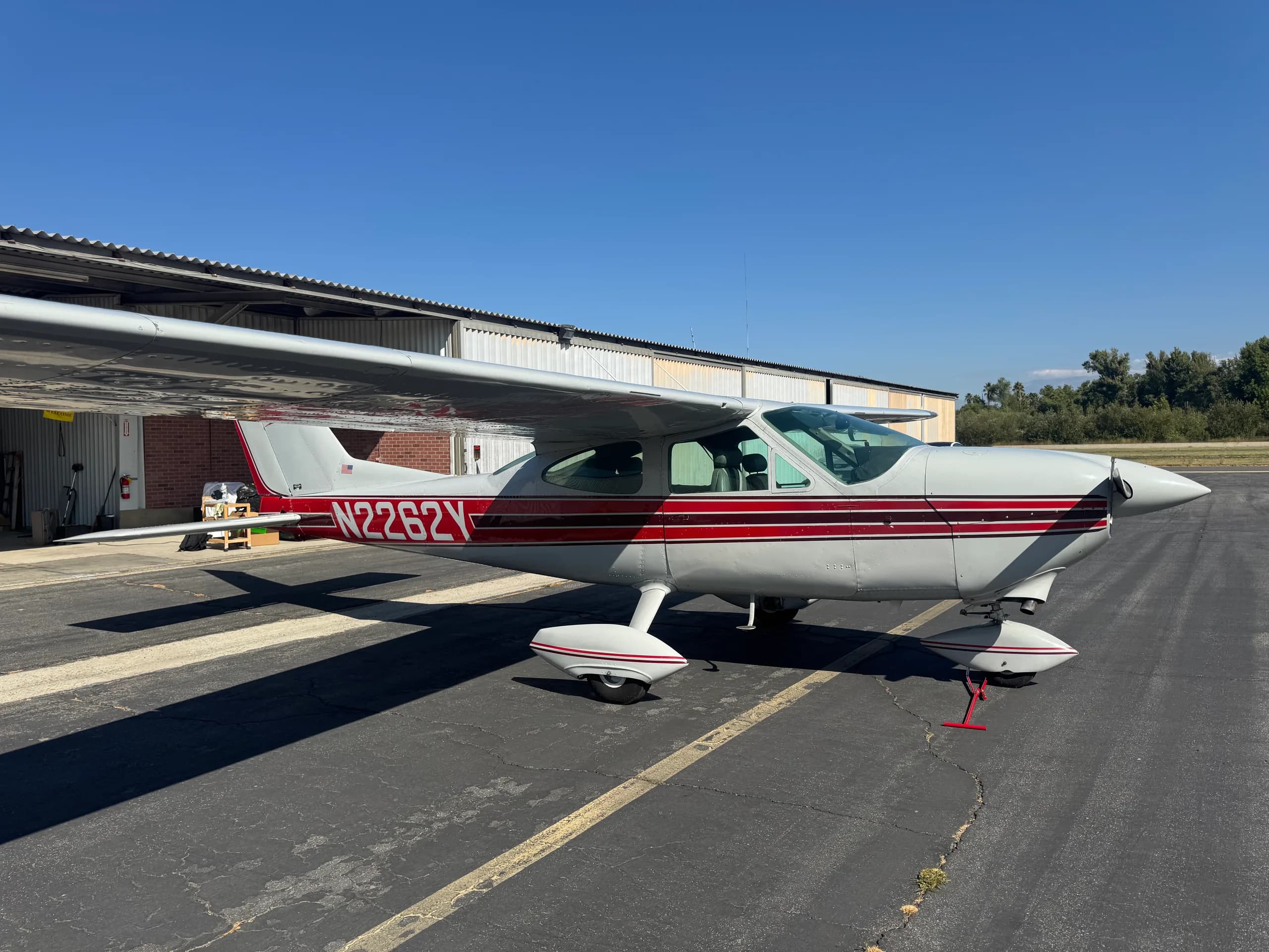 A white and red Cessna N2262Y single-engine aircraft is parked on an asphalt tarmac next to a hangar under a clear blue sky.