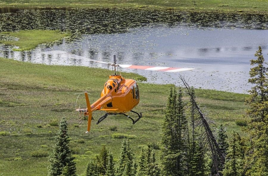 An orange helicopter flying near a lake and trees.
