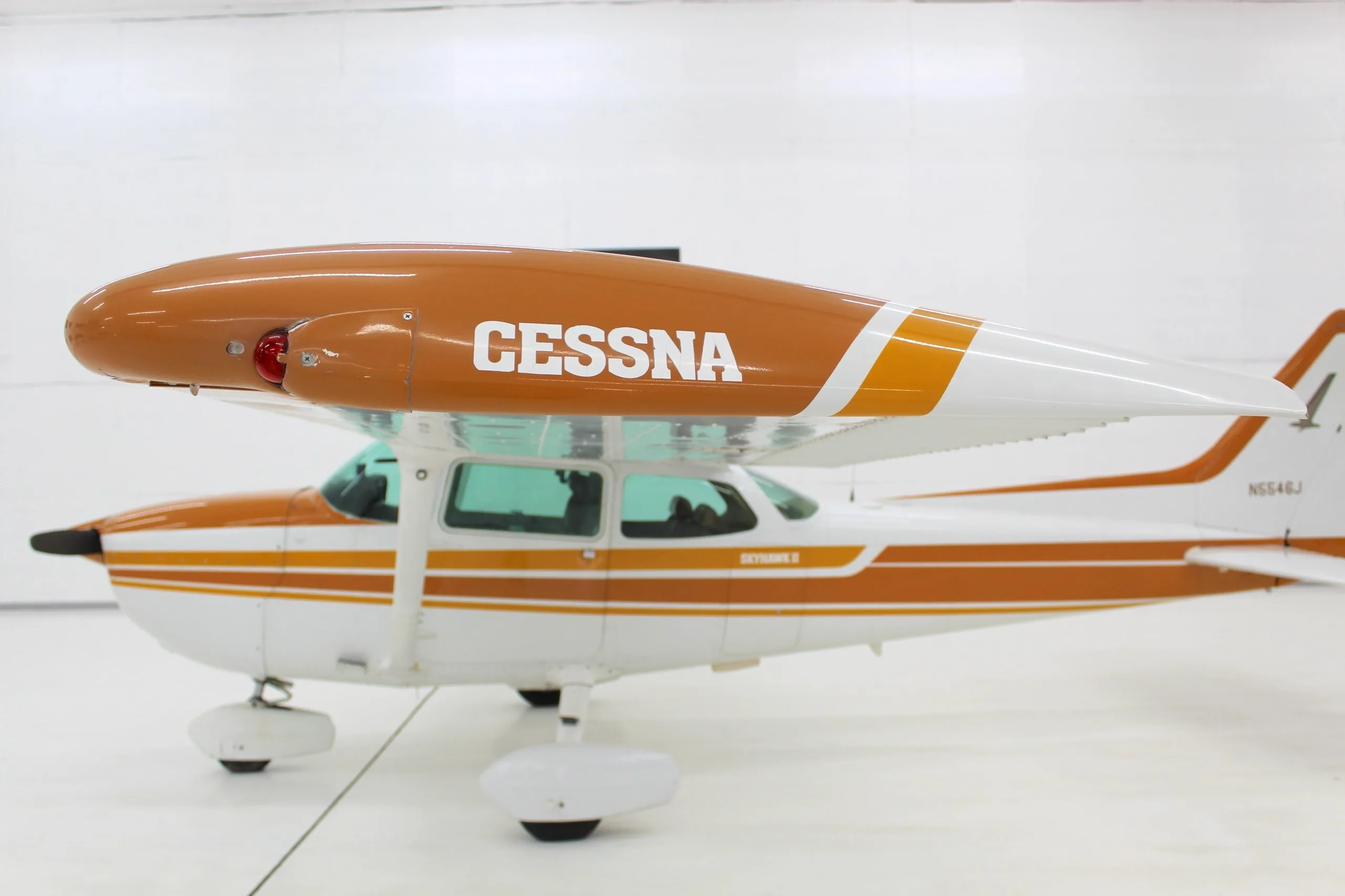 A white and orange Cessna Skyhawk II (N5546J) is parked in a brightly lit hangar, viewed from the side, showing the "CESSNA" text on the wing.
