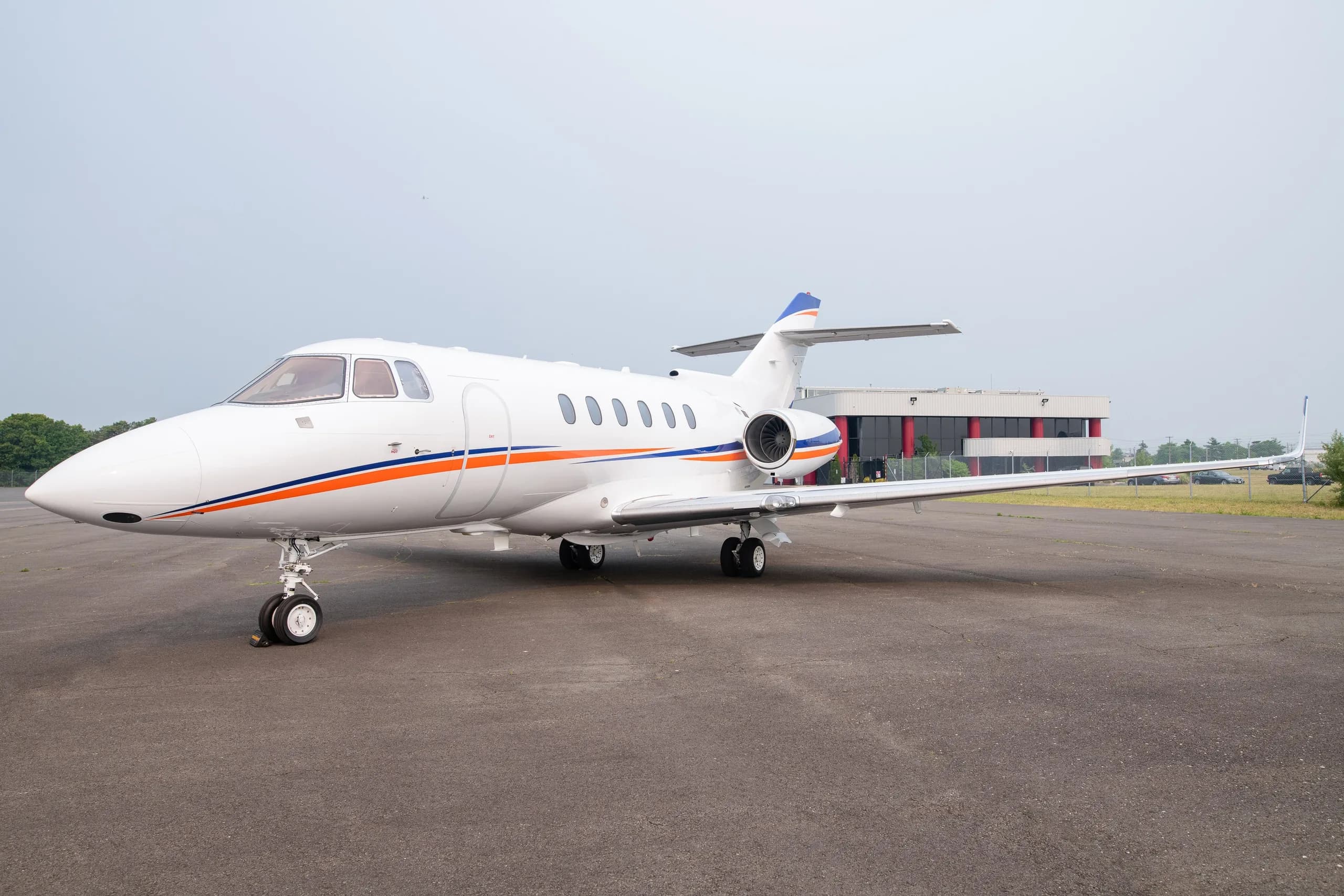 A white business jet with blue and orange stripes on the tarmac, viewed from the front-left.