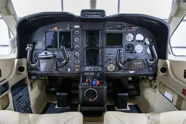 The cockpit of a TBM 700 aircraft, showing the instrument panel with analog gauges and digital displays, and dual yokes.