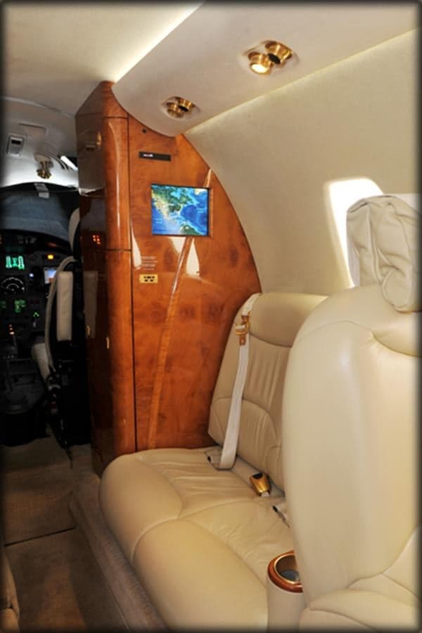 A cream leather passenger seat in a private jet cabin faces a wood-paneled wall with a small display screen showing a map.