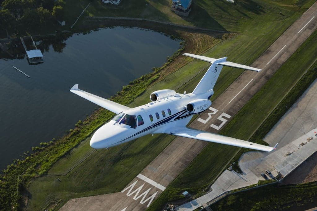A Citation M2 aircraft in flight over a runway and lake.