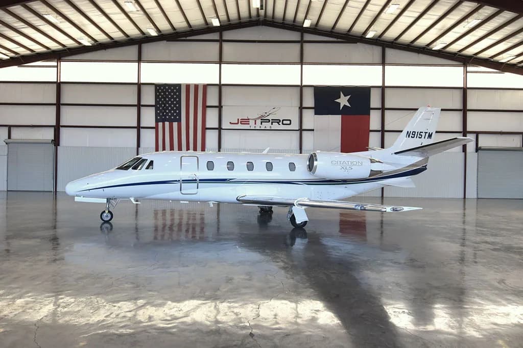 A white Cessna Citation XLS business jet with blue and silver stripes, tail number N915TM, parked inside a hangar with US and Texas flags.