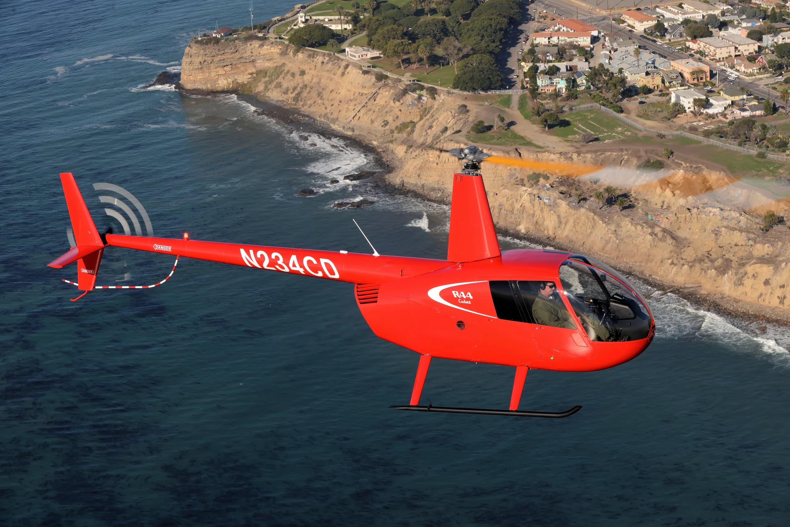 A red Robinson R44 helicopter flying near the coast.