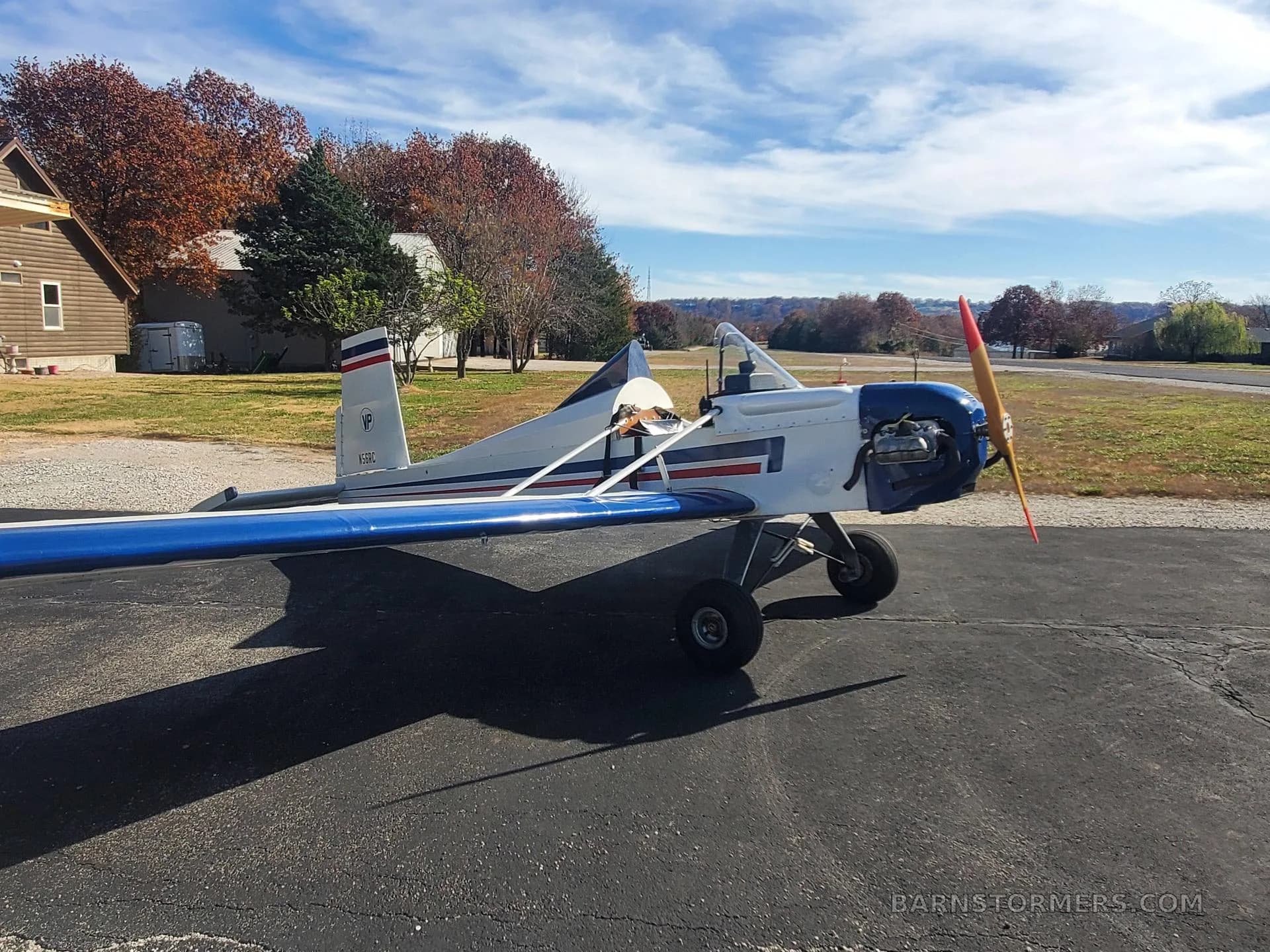 A white and blue single-engine aircraft with a red-tipped propeller is parked on an asphalt surface with autumn trees in the background.