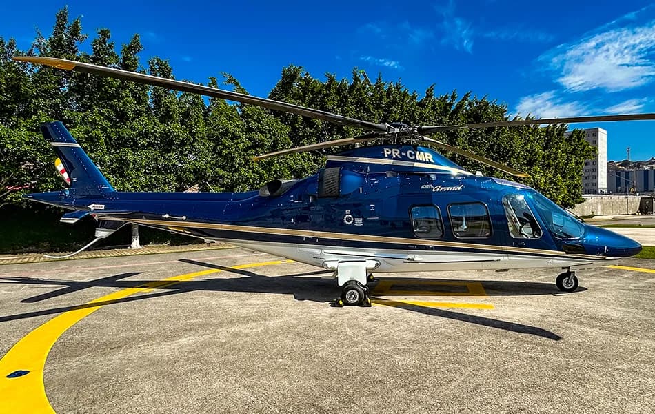 A dark blue and white Agusta Grand helicopter with a gold stripe is parked on a concrete helipad under a clear blue sky.