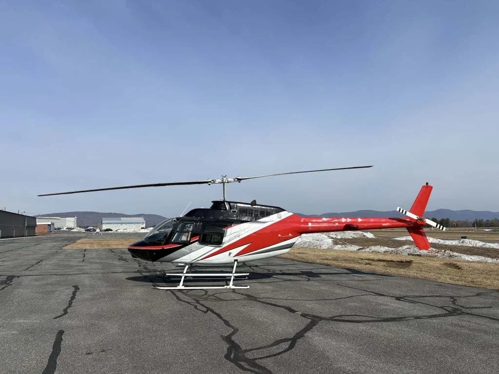 A red, black, and white helicopter with skids on an airfield with some snow patches.