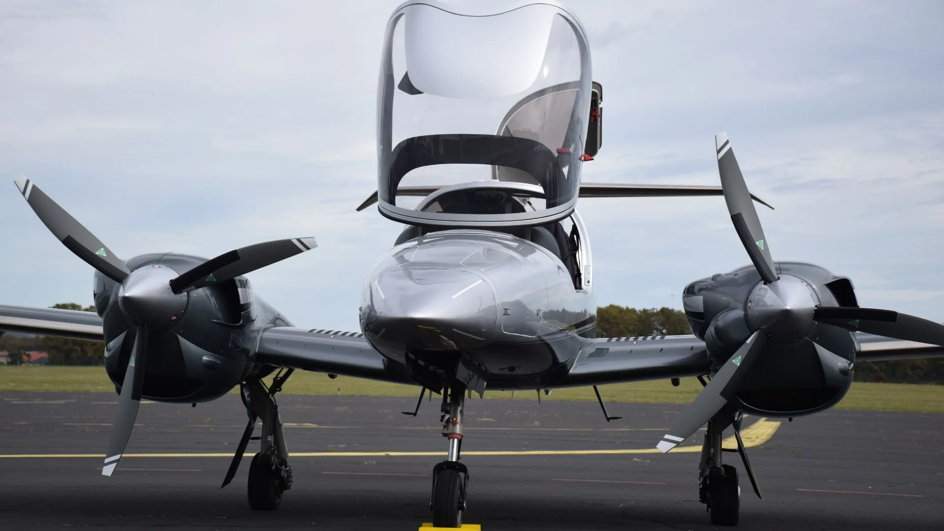 A front view of a silver Diamond DA62 twin-engine aircraft with its canopy open on a tarmac.