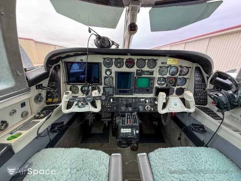 The cockpit of a twin-engine aircraft, featuring a complex instrument panel with analog gauges, two control yokes, and light blue seat covers.