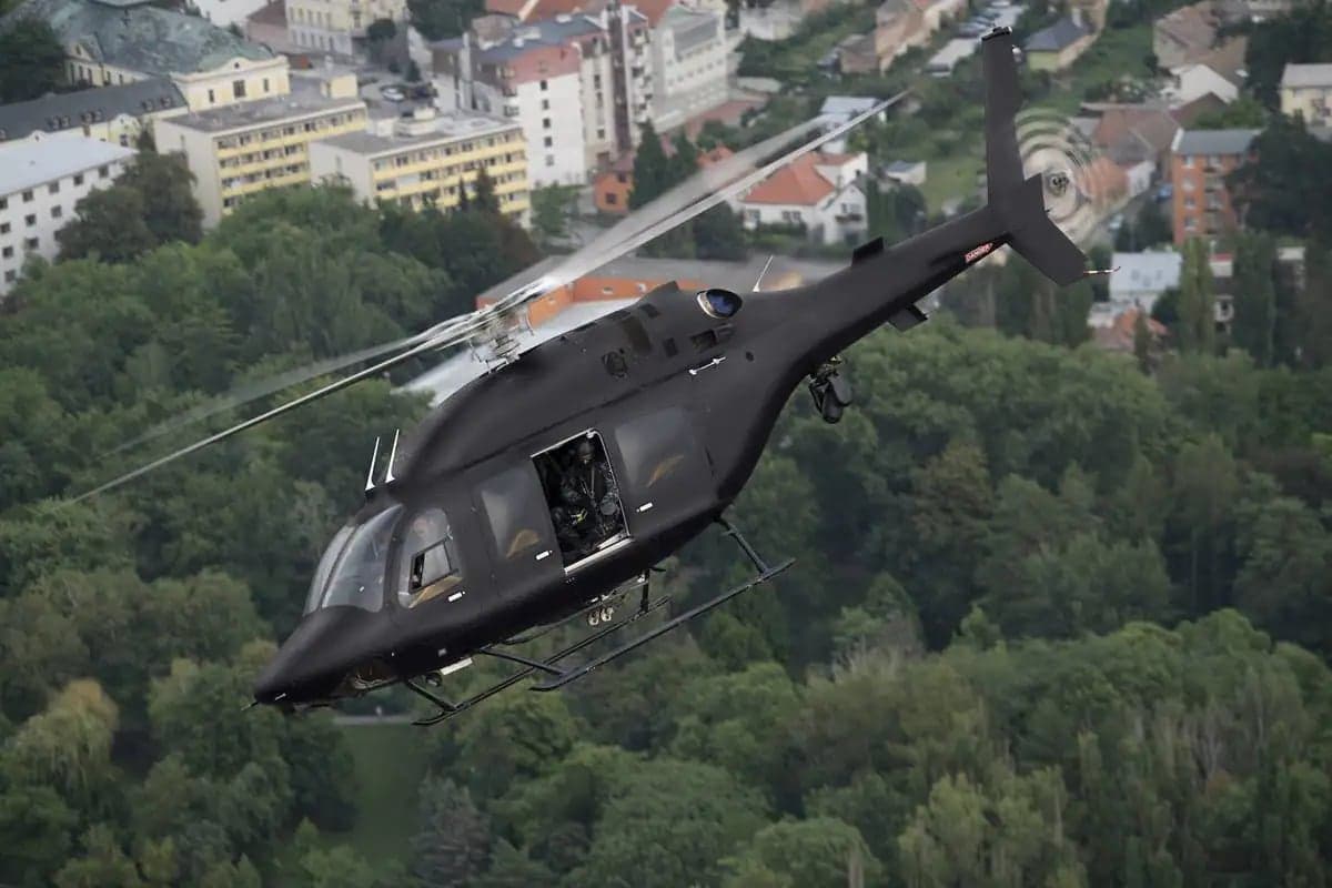 A black helicopter flies over a forest and buildings.