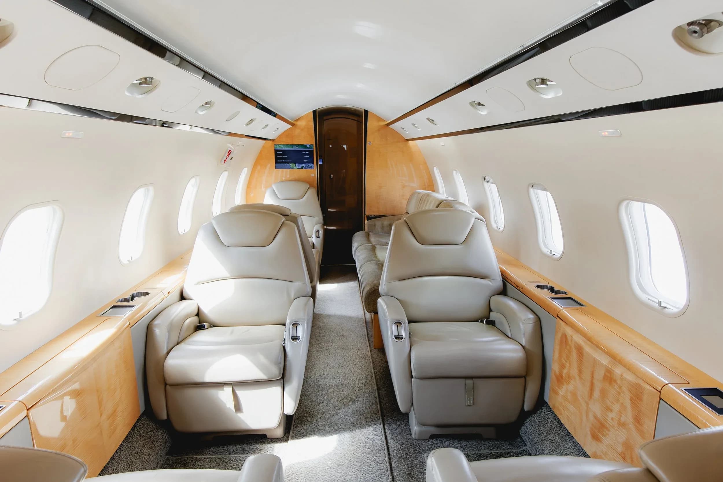 The interior of a private jet cabin with light beige leather seats and light wood paneling, looking towards the rear.