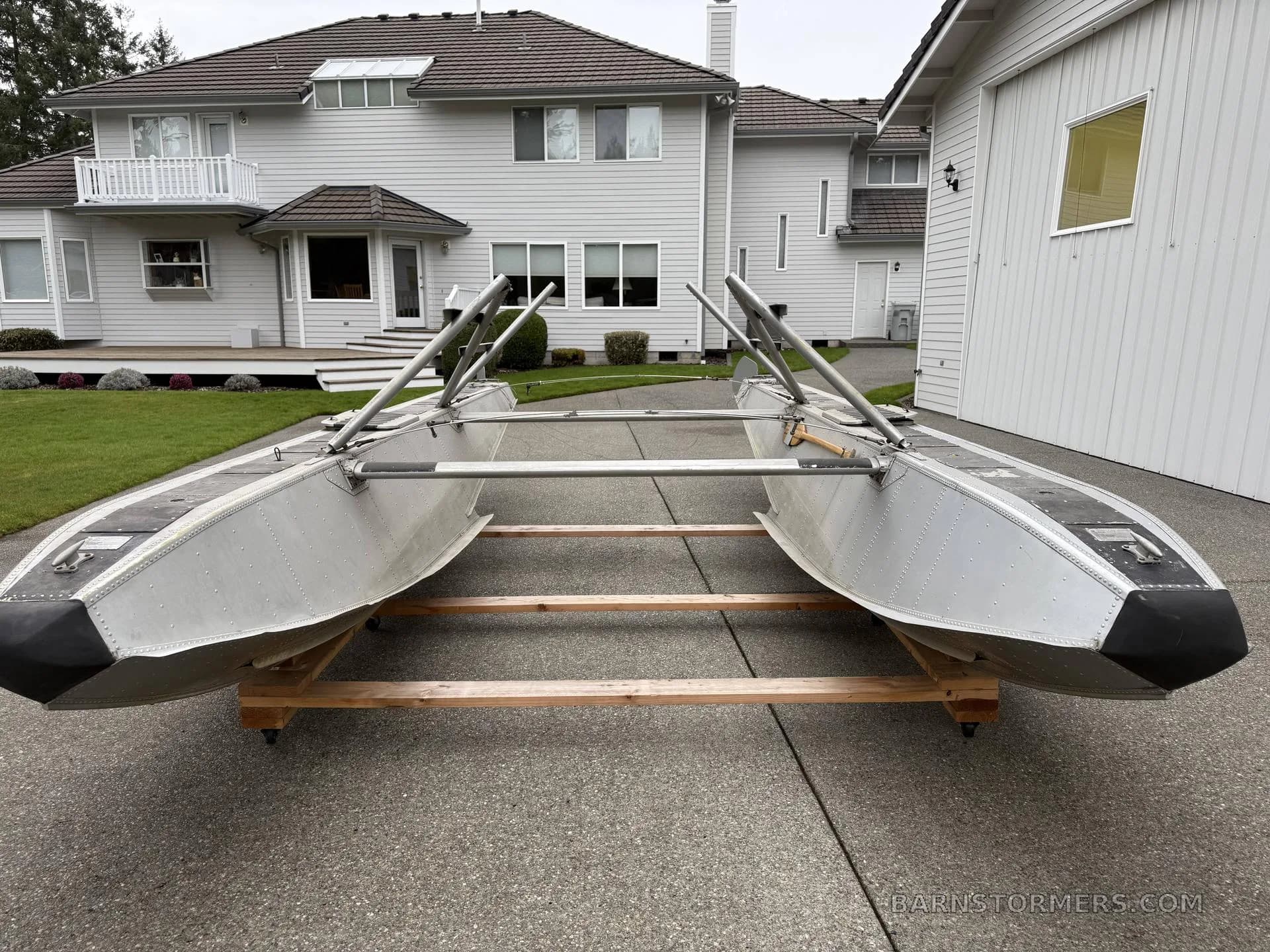 A pair of aluminum aircraft floats with mounting struts, viewed from the front, on a concrete driveway in front of a house.