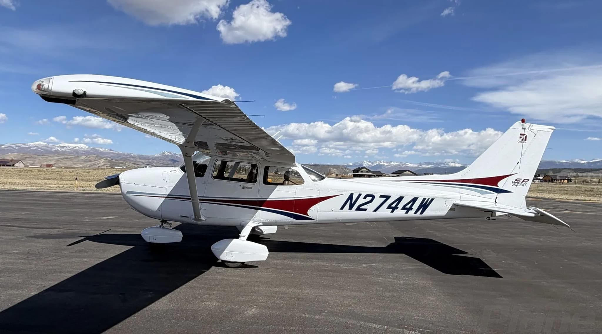 A white Cessna 172 Skyhawk with red and blue stripes, registration N2744W, parked on an airfield with mountains in the background.