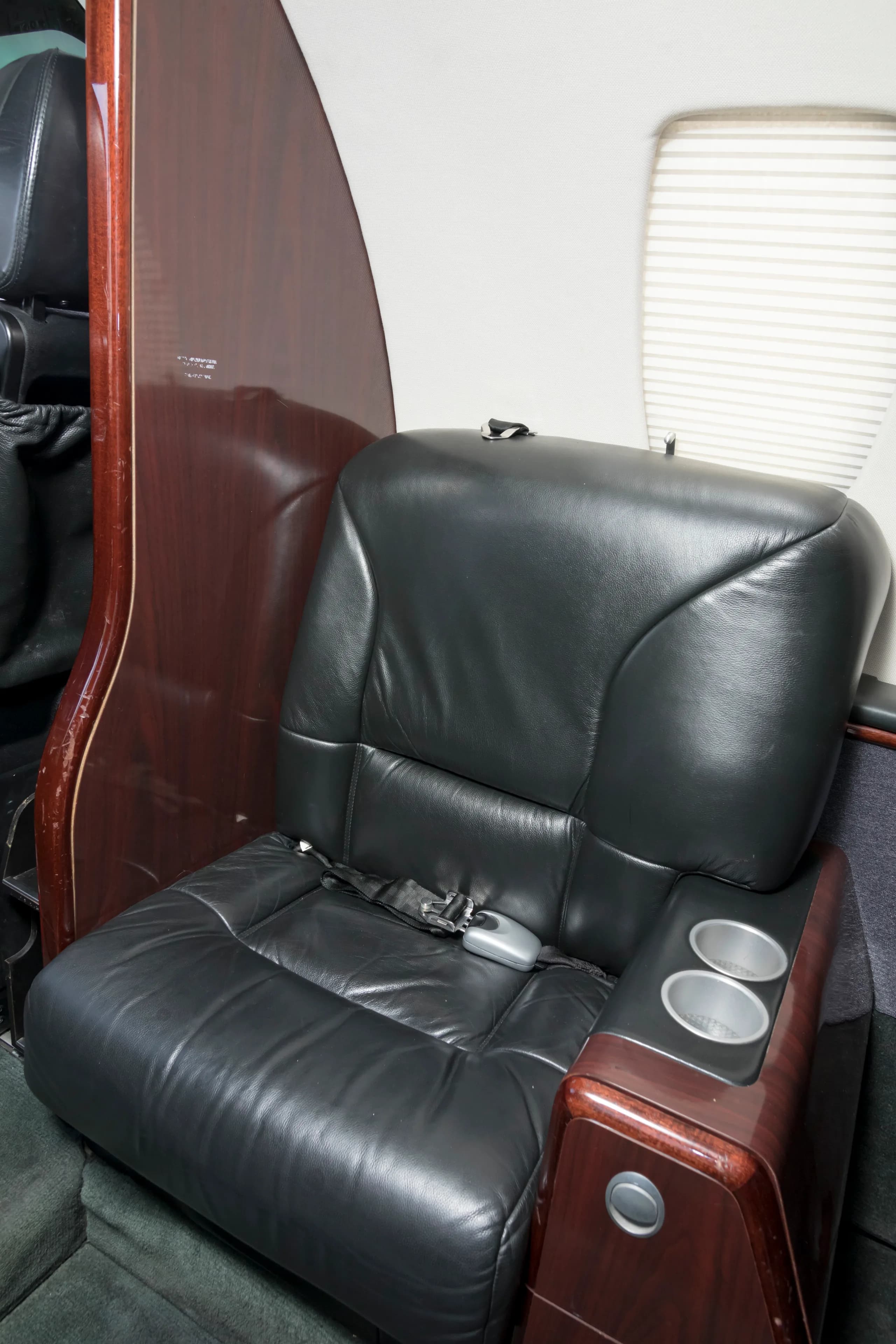 A close-up of a black leather passenger seat in a private jet cabin, with a dark wood panel and cup holders.