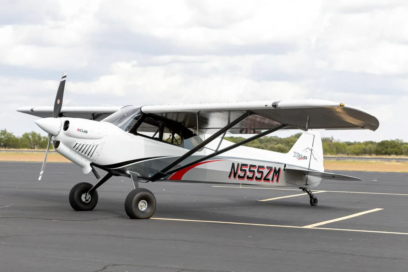 A white and grey X-Cub aircraft with red accents and registration N555ZM parked on an asphalt runway under a cloudy sky.
