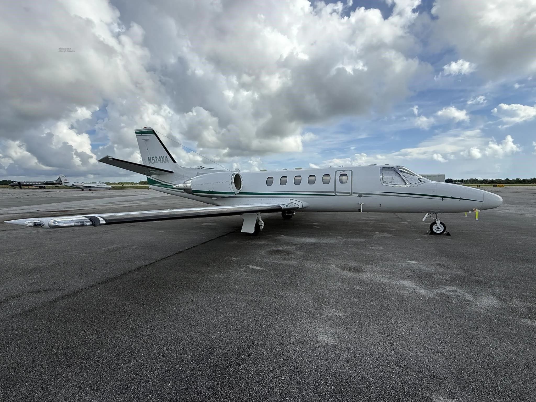 A white business jet N524XA with green stripes, parked on an airport tarmac with other aircraft in the distance.