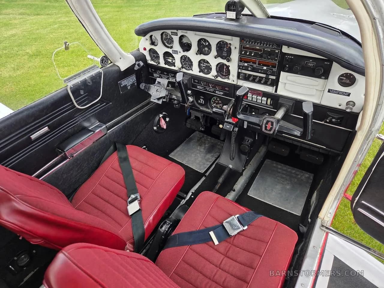 The cockpit of a Piper Cherokee aircraft features a black instrument panel, dual yokes, and red fabric seats.