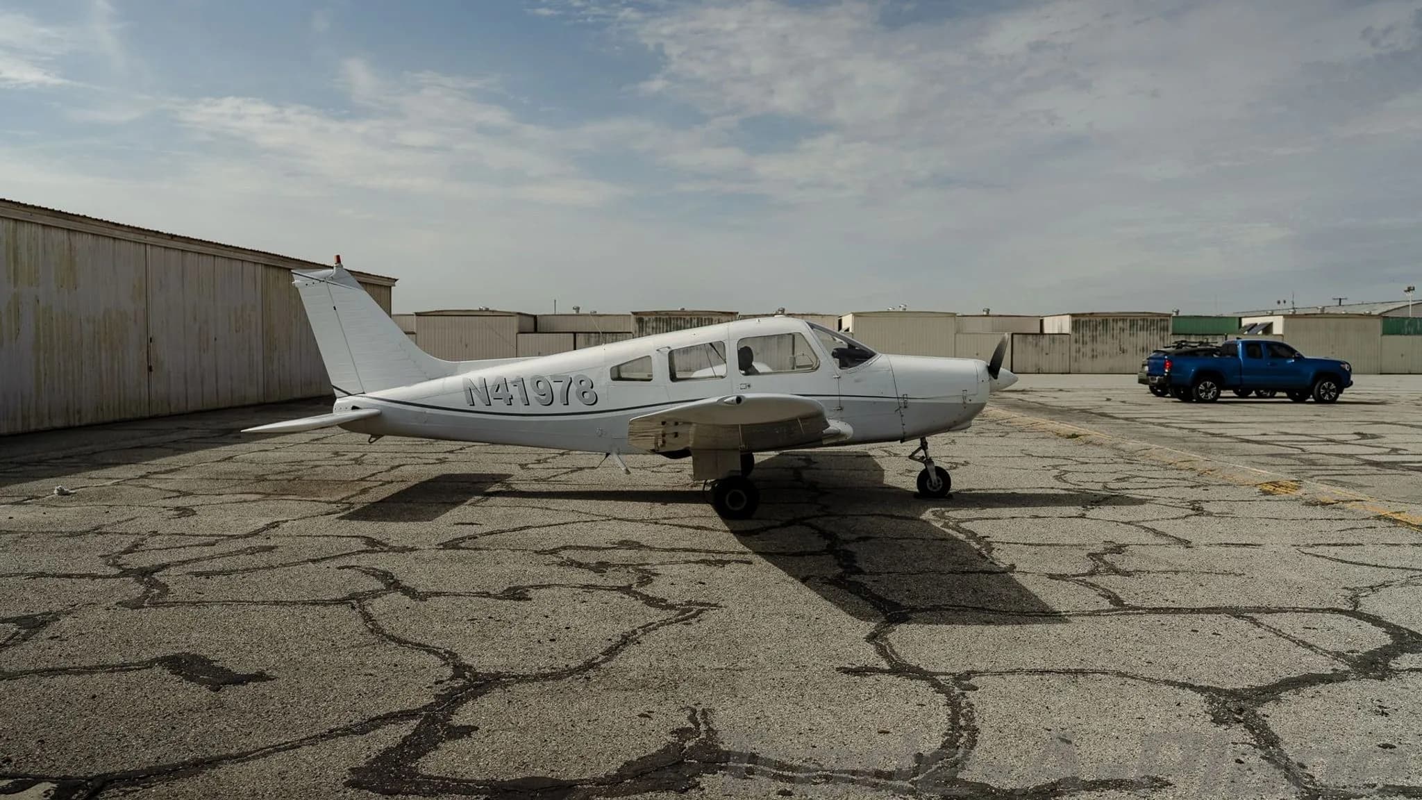 A white Piper Cherokee N41978 parked on a cracked tarmac next to a blue truck.