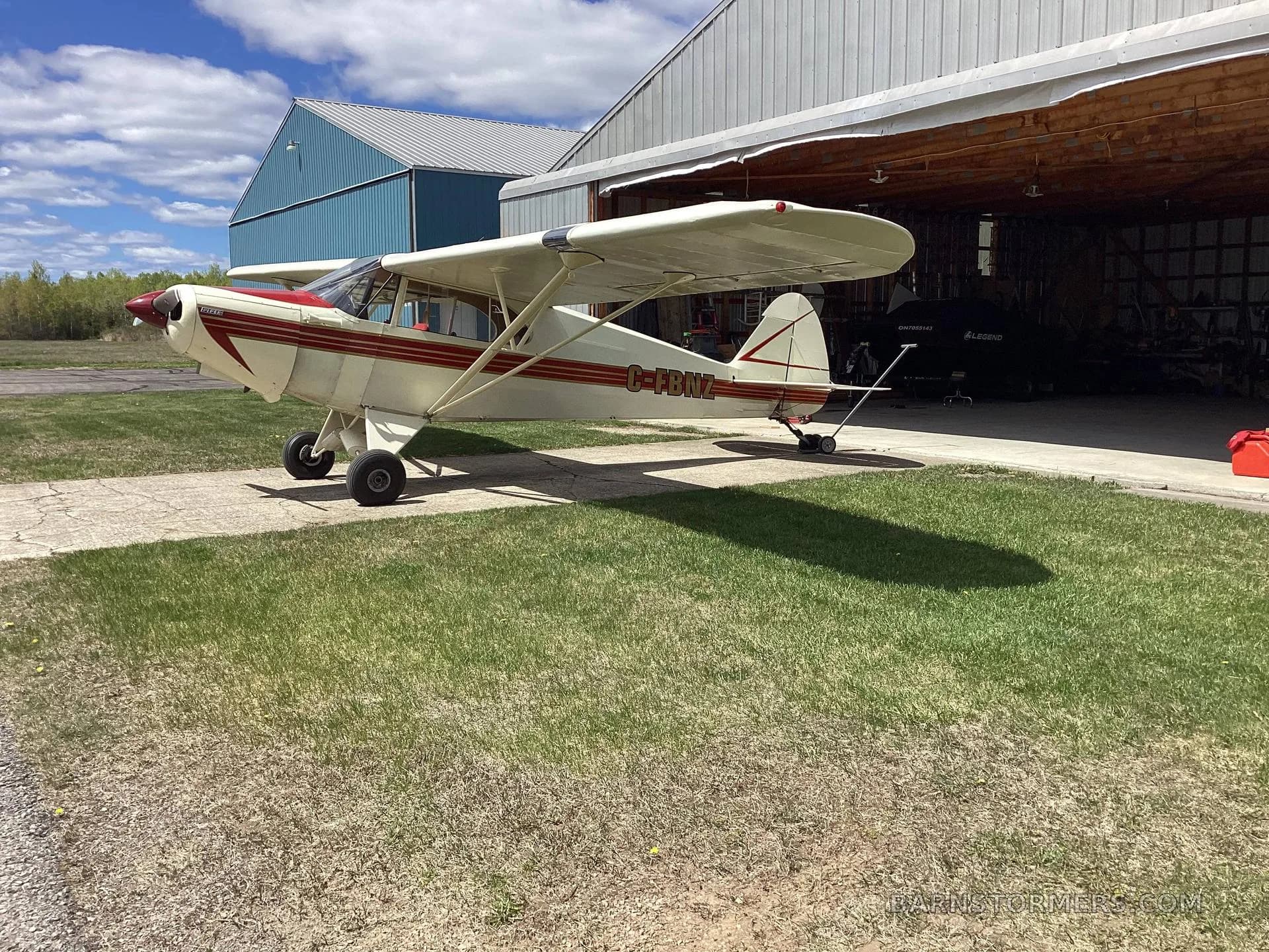 A cream and red high-wing aircraft with tail number C-FBNZ parked on a concrete pad outside a hangar, viewed from the rear-right.