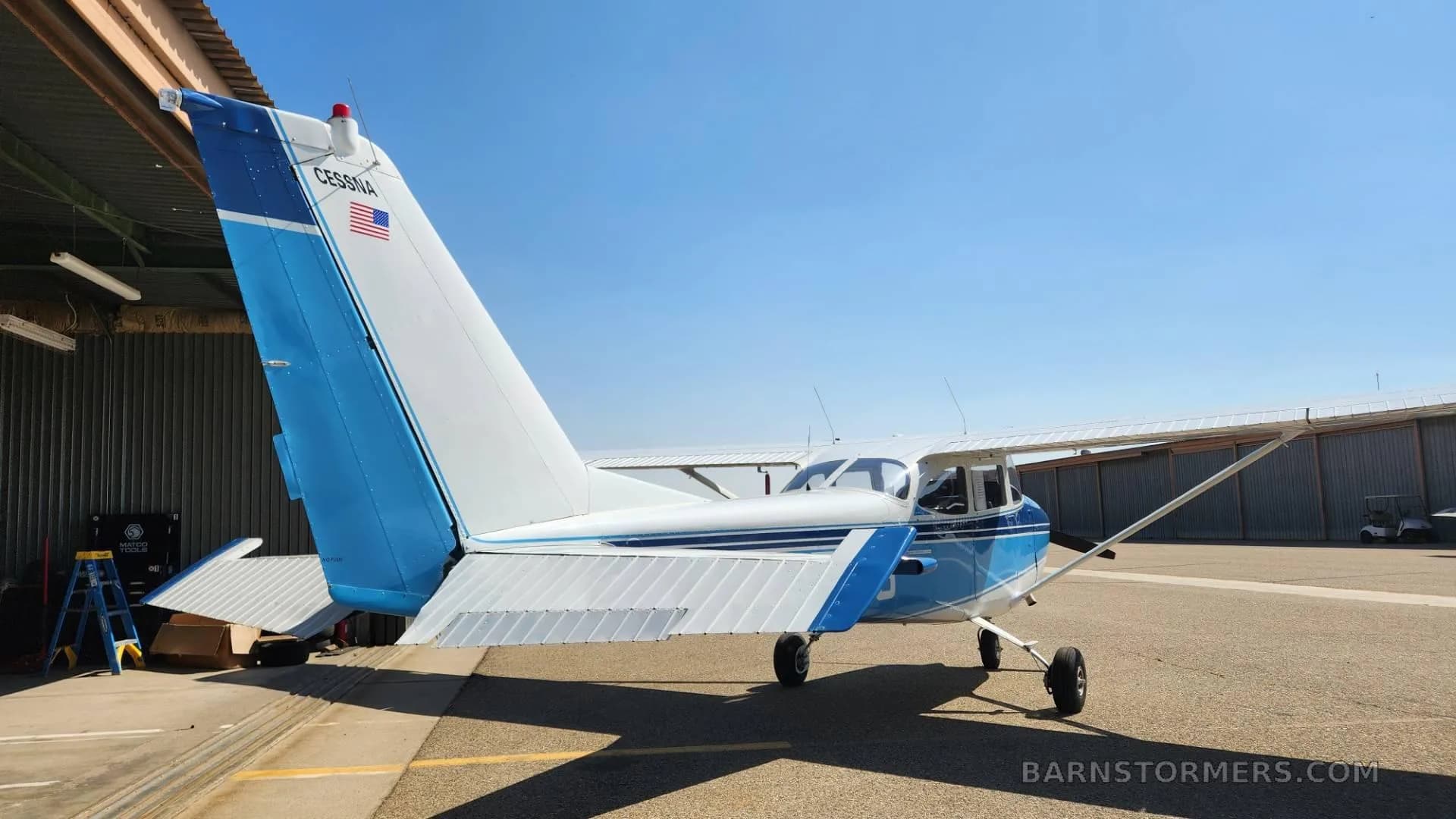 A blue and white Cessna aircraft is parked on an airfield, viewed from the rear-right, with a hangar in the background.