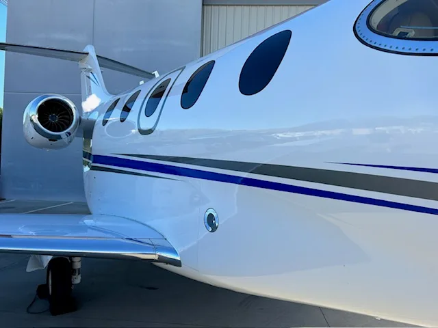 A close-up view of the white fuselage of a business jet with blue and grey stripes, showing the cabin windows and an engine.