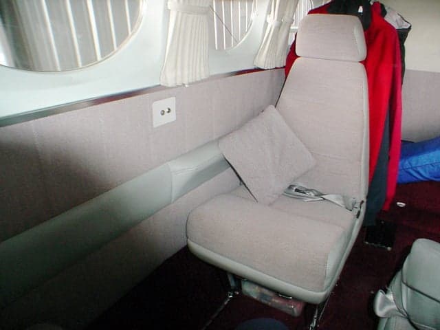 The aircraft cabin interior with a single beige fabric seat, a matching pillow, and pleated window shades.