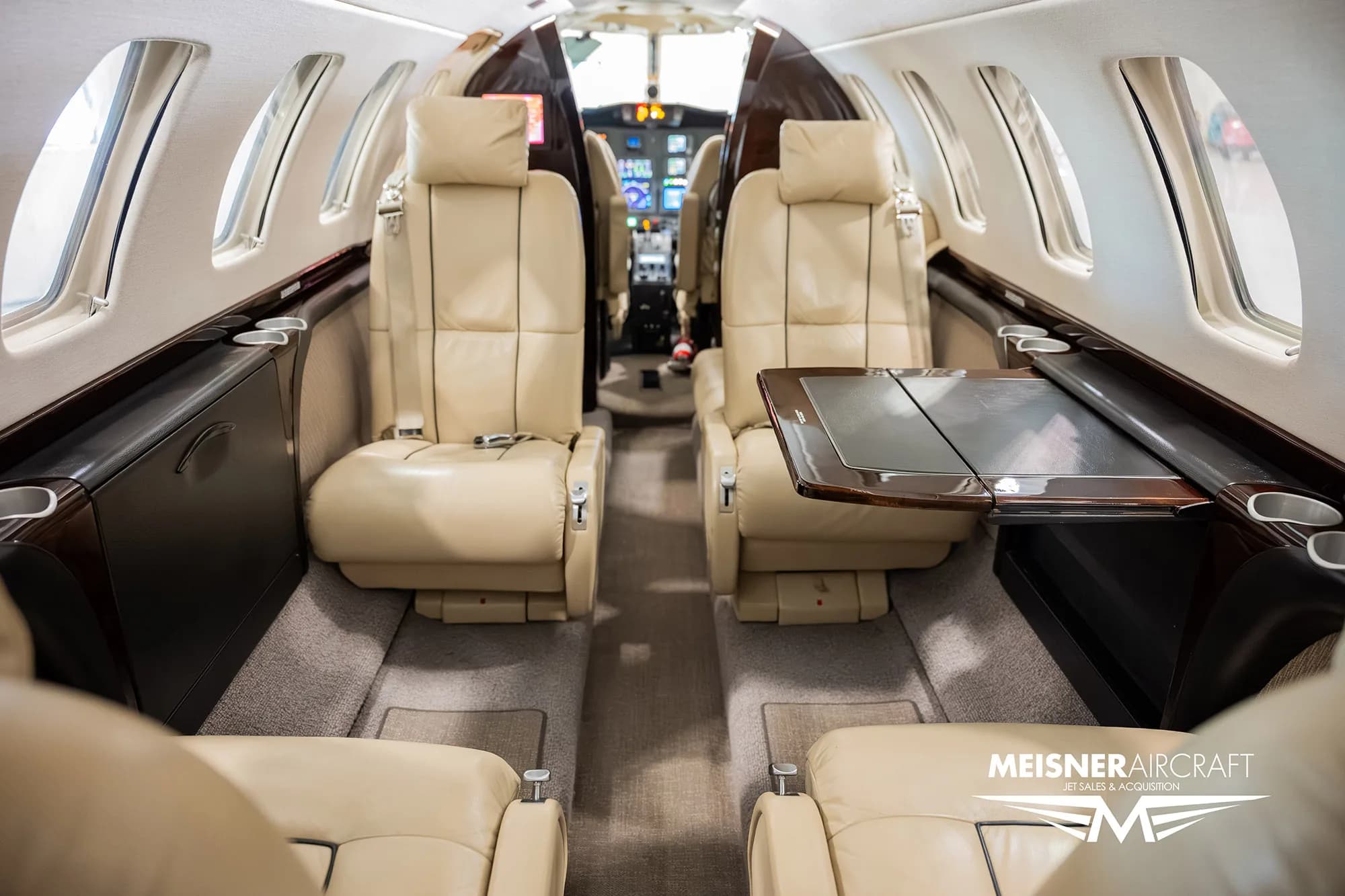 The interior cabin of a business jet, featuring beige leather seats and a small wooden table deployed between them.