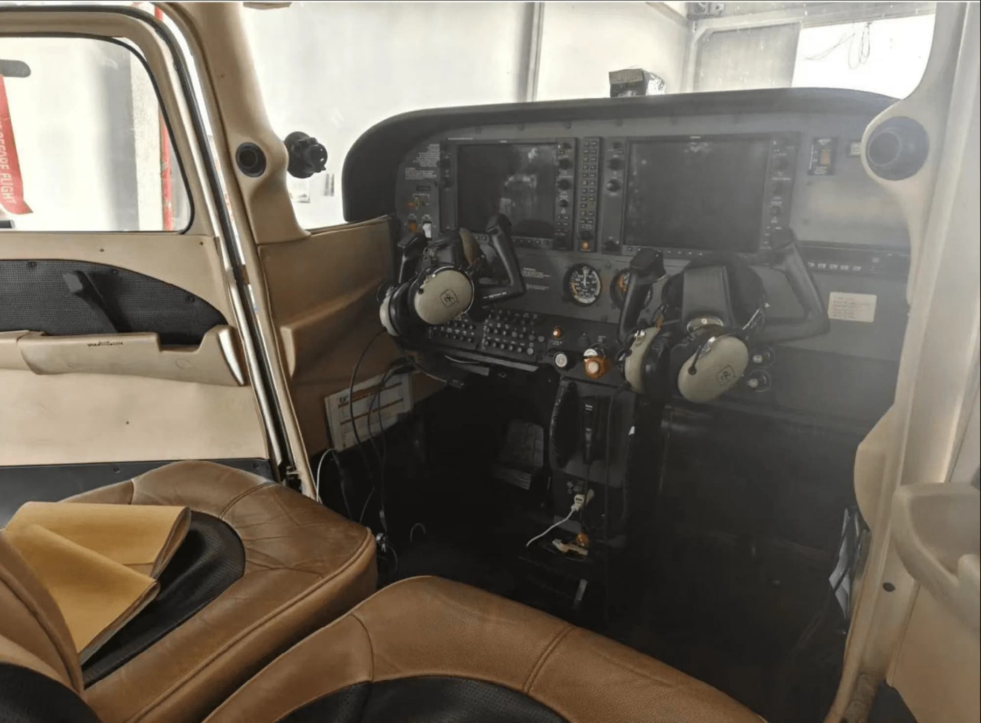 The cockpit of a small aircraft showing the instrument panel with multiple screens, yokes, and tan leather seats.