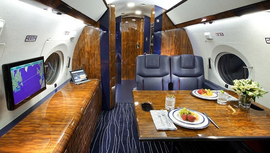 The private jet's cabin interior with a dining table set for two, an entertainment screen, and polished dark wood paneling.