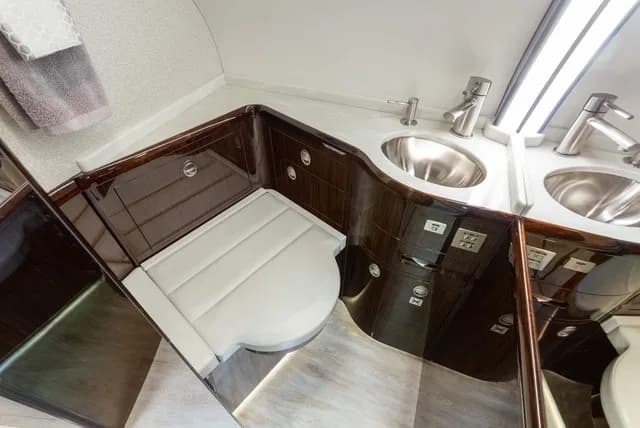 A private jet lavatory with dual sinks, a toilet, and dark wood cabinetry.