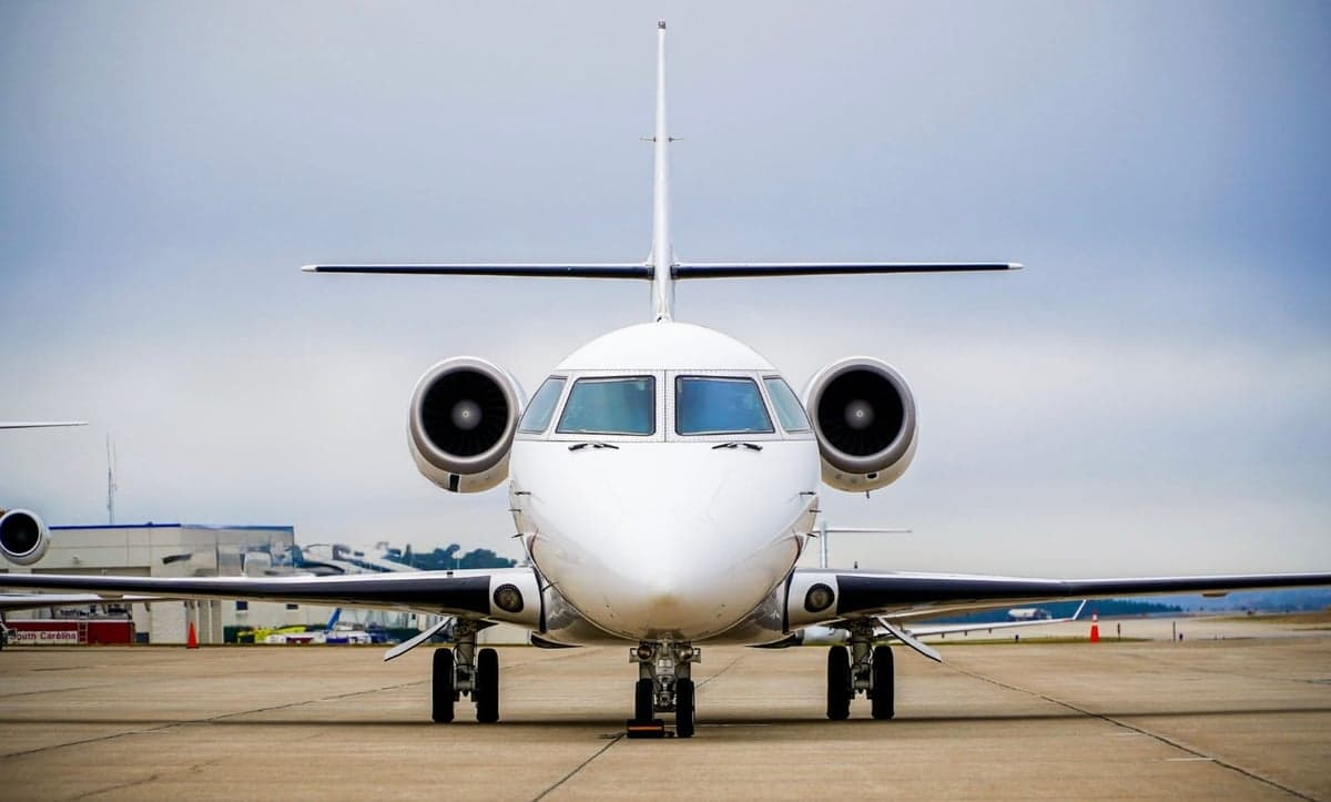 A white private jet with two rear-mounted engines is seen from a head-on view, parked on an airport tarmac under a cloudy sky.