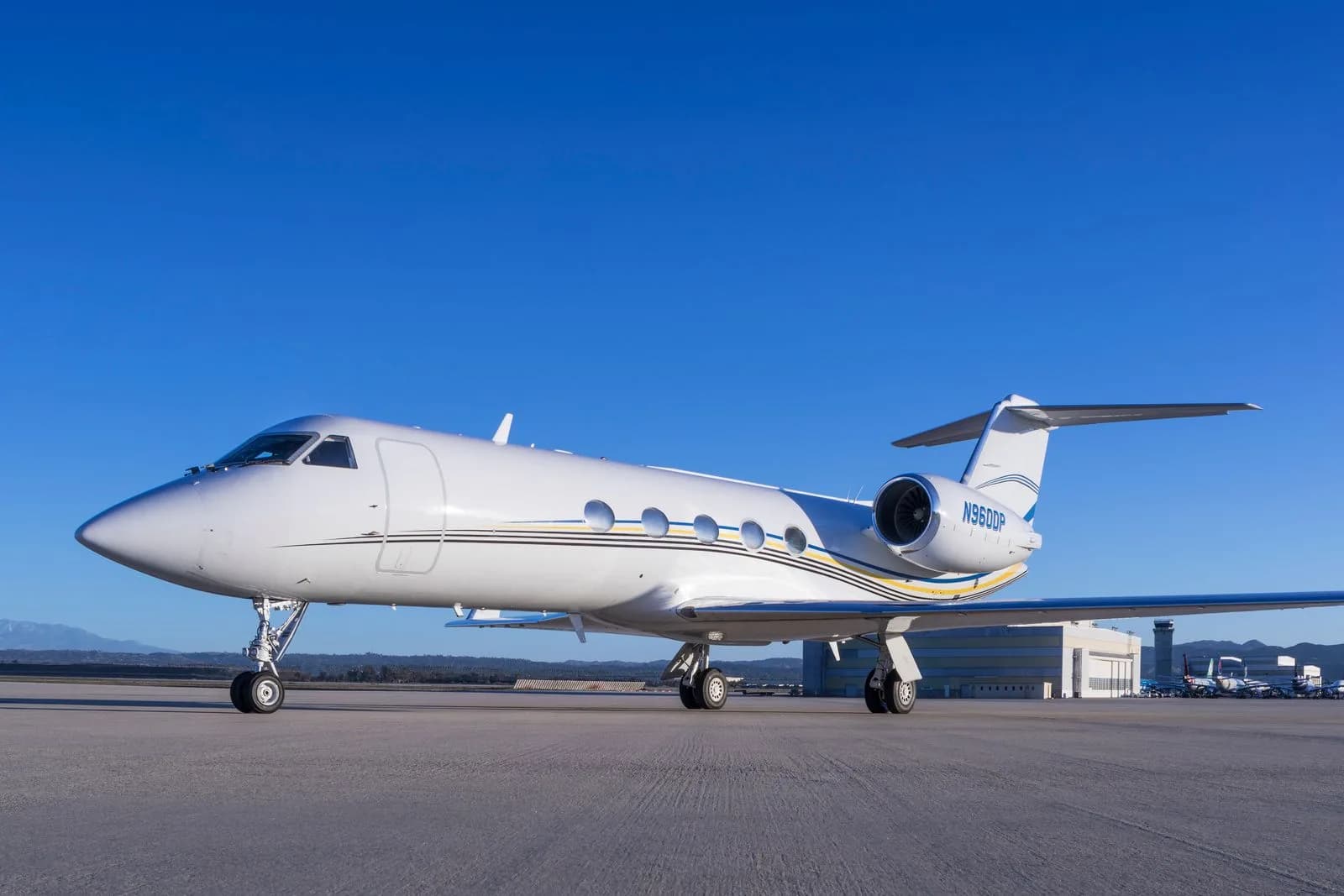 A white Gulfstream business jet with black, yellow, and blue stripes is parked on an airport tarmac under a clear blue sky.
