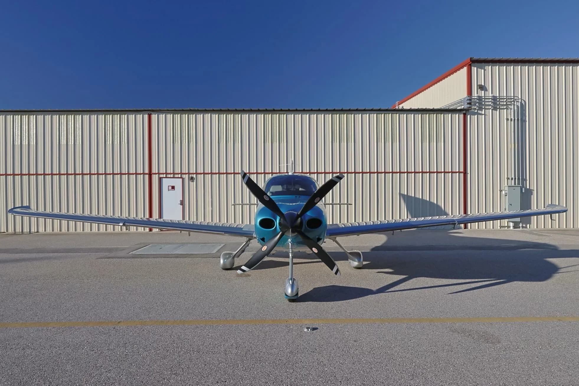 A head-on view of a teal and silver Cirrus SR22T GTS aircraft (N818PK) parked in front of a hangar.