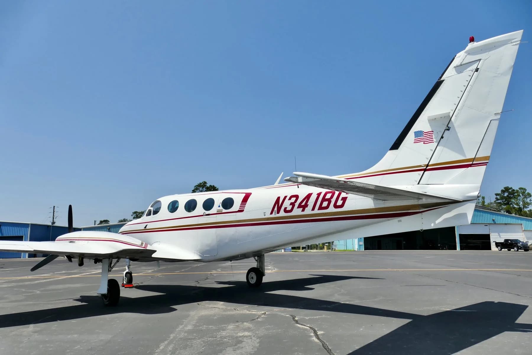 Rear three-quarter view of a white twin-engine propeller aircraft N341BG parked on an airfield next to a hangar.