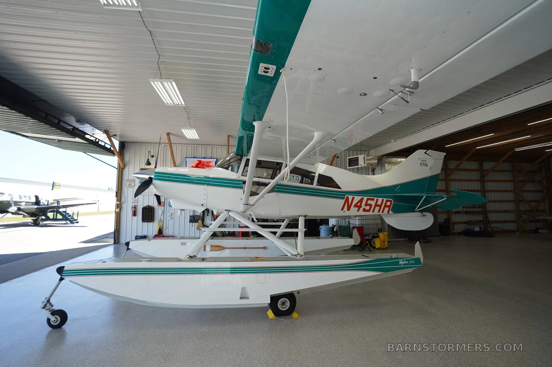 A white and teal floatplane, registration N45HR, is parked inside a hangar, viewed from the front-right.