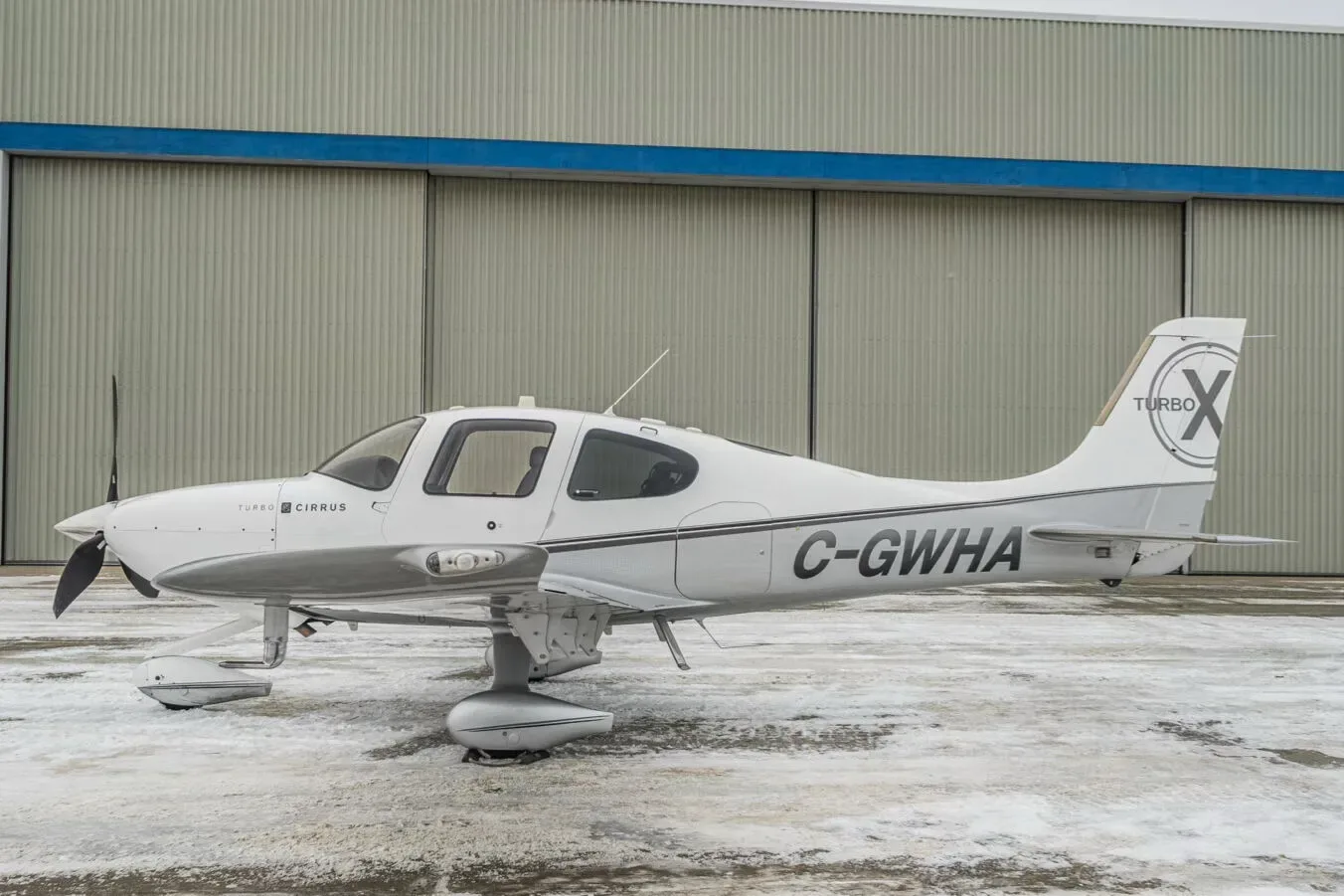 A white and grey Cirrus SR22T aircraft (C-GWHA) with a 'Turbo X' logo on the tail, parked on a snowy ground in front of a hangar.